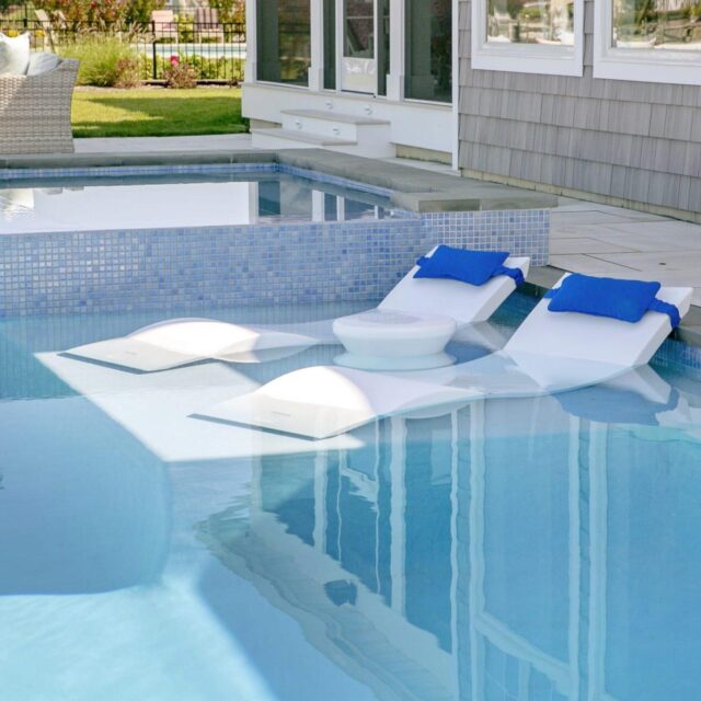 Two white pool loungers with blue pillows are partially submerged in a luxury pool beside a custom spa, creating the perfect backyard outdoor living escape.