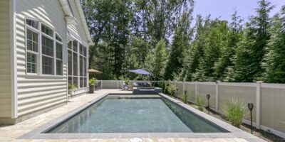 Rectangular backyard luxury pool beside a house, surrounded by a stone patio, tall trees, white fence, and patio furniture with a blue umbrella in the background.
