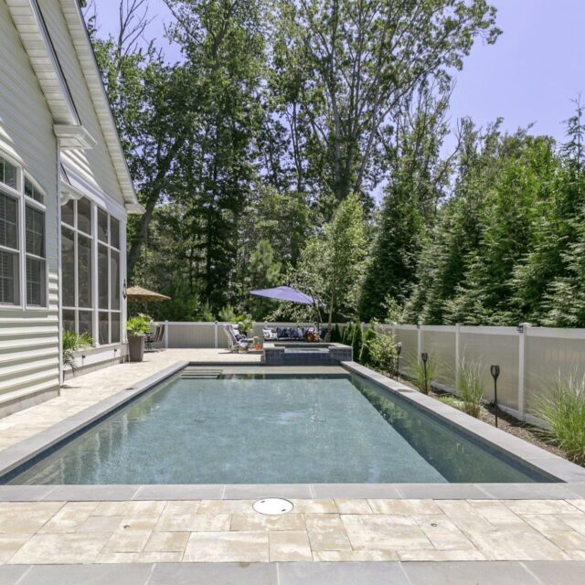 Rectangular backyard luxury pool beside a house, surrounded by a stone patio, tall trees, white fence, and patio furniture with a blue umbrella in the background.