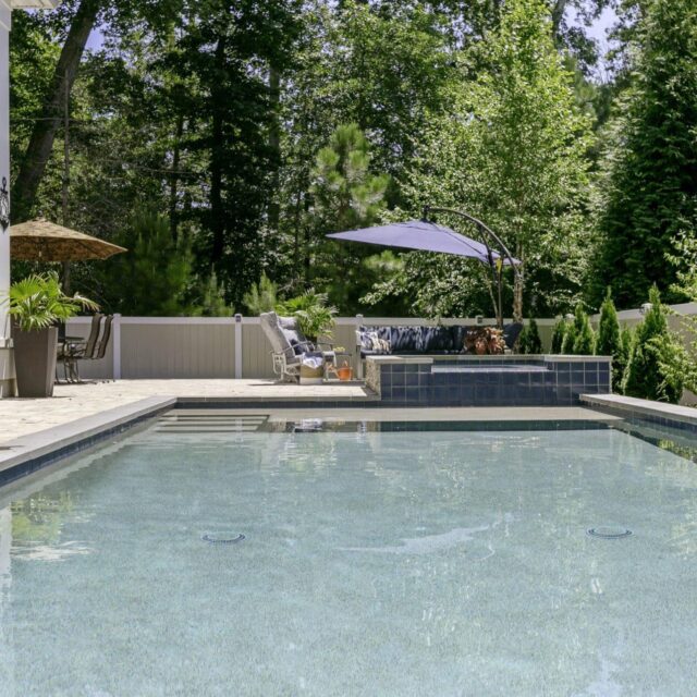 Rectangular inground pool with clear water, surrounded by trees, a patio area, outdoor furniture, and a shaded seating area in the background—perfect for enjoying luxury pools at home.