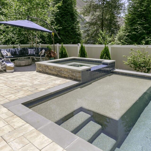 A backyard patio designed for outdoor living features an inground pool with steps, a custom spa hot tub, cushioned seating area with fire pit and umbrella, all bordered by a fence and tall greenery.