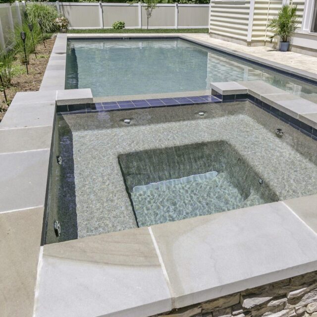 Rectangular hot tub with jets connected to a swimming pool in a backyard, surrounded by stone and tile decking—perfect for those seeking elegant inground pools and custom spas.