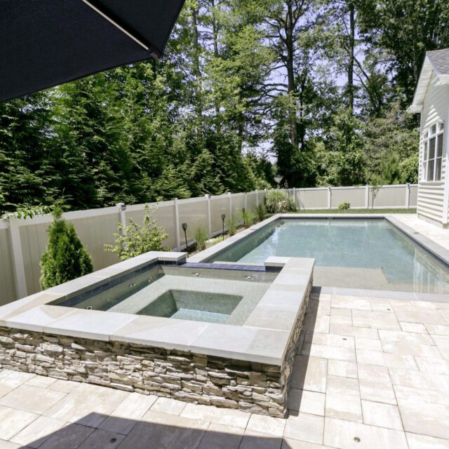 A rectangular backyard luxury pool with an attached stone hot tub, surrounded by a tiled patio and white privacy fence, bordered by trees and shrubs—perfect for outdoor living.