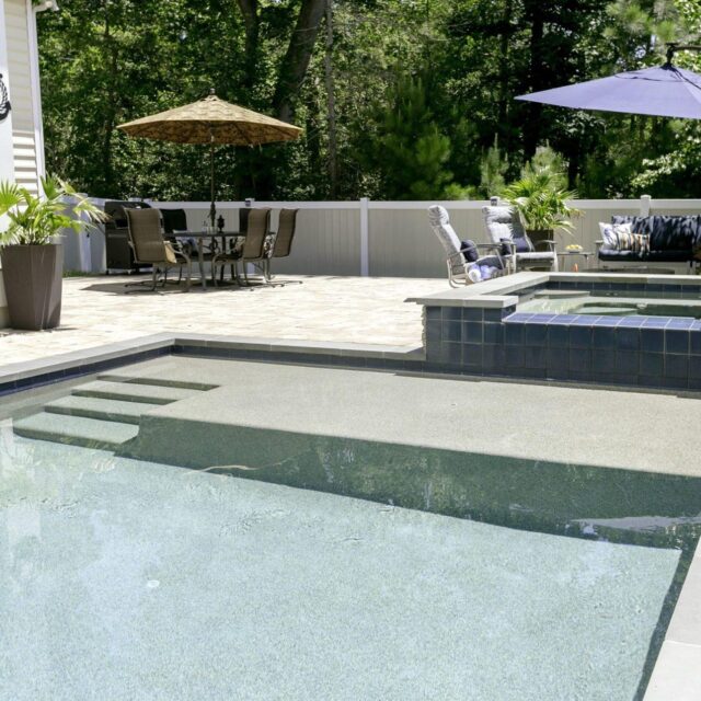 A backyard showcases luxury pools with a connected hot tub, patio seating under umbrellas, potted plants, and is bordered by a white fence and trees—perfect for stylish outdoor living.