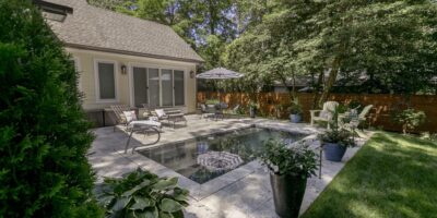 Backyard with a rectangular inground pool, lounge chairs, potted plants, umbrella, and a grassy lawn bordered by trees and a wood fence—perfect for enjoying the benefits of modern fiberglass pools.