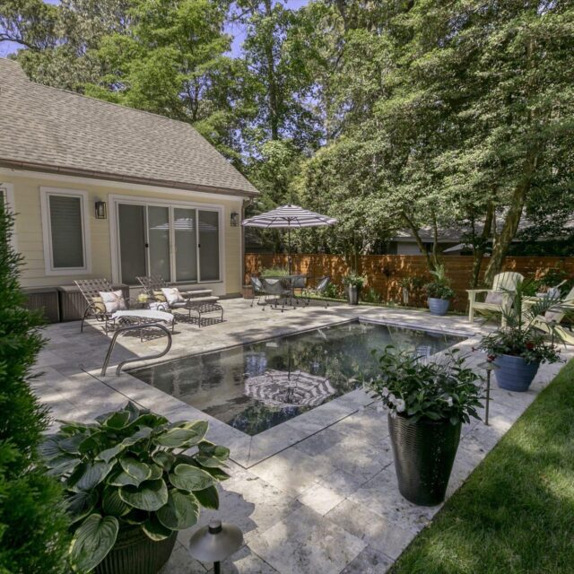 Backyard with a rectangular inground pool, lounge chairs, potted plants, umbrella, and a grassy lawn bordered by trees and a wood fence—perfect for enjoying the benefits of modern fiberglass pools.