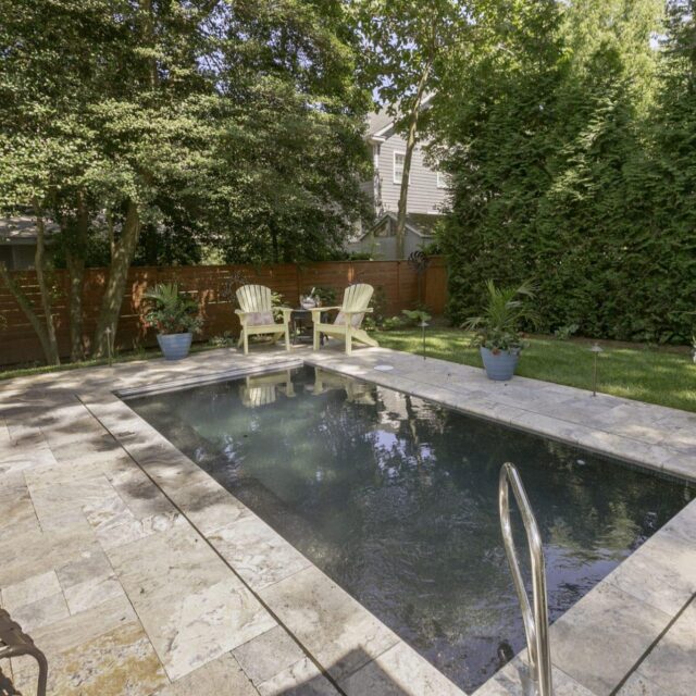 Rectangular inground pool surrounded by stone tiles, two yellow chairs, potted plants, and trees providing partial shade—perfect for outdoor living.