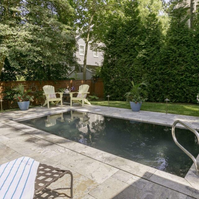 A small rectangular backyard luxury pool with a ladder, surrounded by stone patio, lounge chairs, potted plants, and two Adirondack chairs with a table in a shaded garden setting.