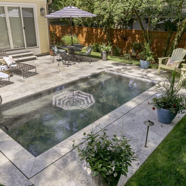 A small rectangular luxury pool surrounded by stone tiles, lounge chairs, plants, and a patio table with an umbrella.