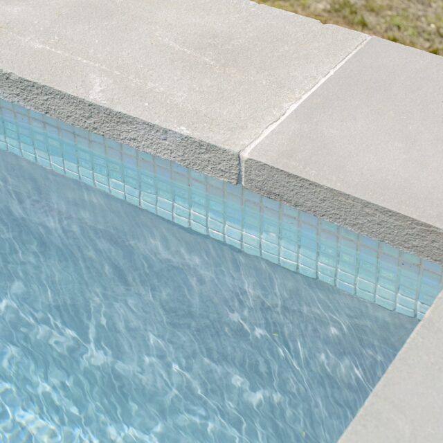 Close-up view of an inground pool corner showing clear blue water, light blue square tiles, and concrete pool edging with grass visible in the background.
