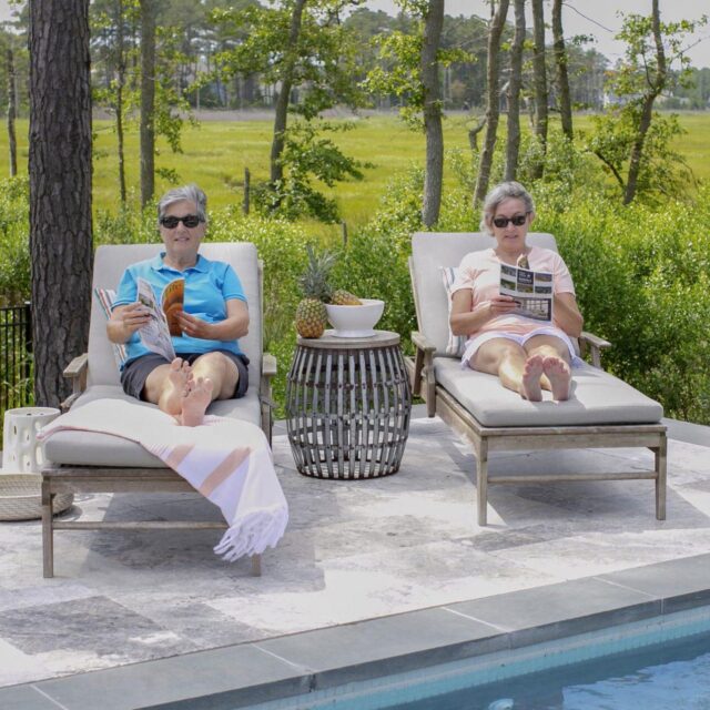 Two people relax on poolside lounge chairs by a luxury pool, each reading a book, surrounded by greenery and trees in the background.
