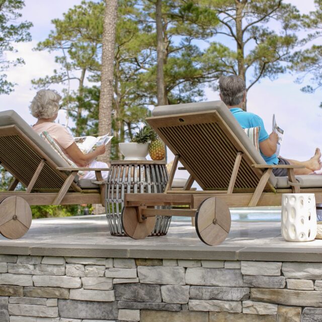 Two older adults sit on wooden lounge chairs by a fiberglass pool, reading and relaxing, with trees, sky, and a glimpse of outdoor kitchens in the background.