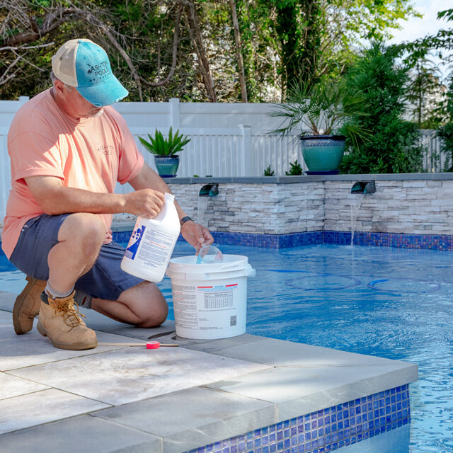 A person kneels by a swimming pool, pouring pool chemicals from a container into a bucket, with lush plants and a white fence in the background—an essential step for maintaining sparkling luxury pools.