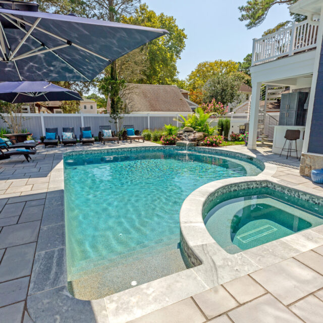 A backyard with a rectangular swimming pool and custom spa, surrounded by lounge chairs, umbrellas, lush landscaping, and space for outdoor living next to a blue house.