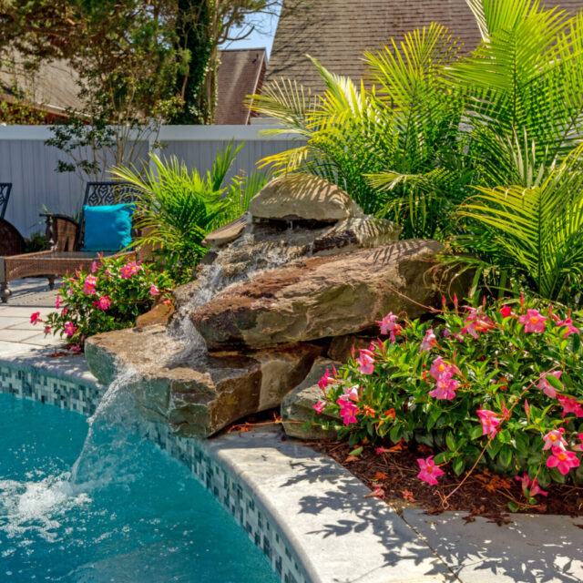 A stone waterfall feature pours into an inground pool, surrounded by tropical plants and pink flowers, with lounge chairs in the background—perfect for relaxing outdoor living.