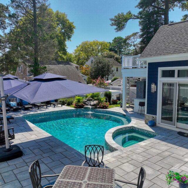 A backyard with a luxury pool, custom spa, lounge chairs under large umbrellas, and a patio table next to a blue house on a sunny day.