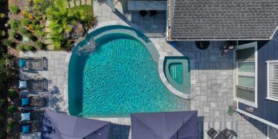 Aerial view of a backyard featuring a luxury pool with an attached custom spa, patio furniture, lounge chairs, umbrellas, and beautiful landscaping.