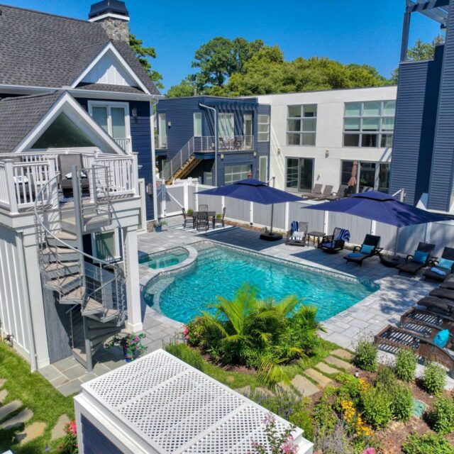 A modern house with a fenced backyard featuring a luxury pool, lounge chairs, umbrellas, landscaped garden, outdoor kitchen, and a small balcony overlooking the pool area.