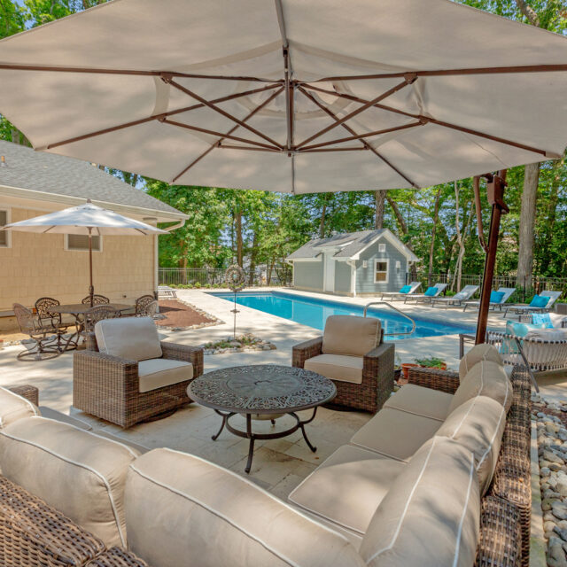 Outdoor patio area with cushioned wicker furniture under a large umbrella, overlooking luxury pools and lounge chairs in a backyard setting.