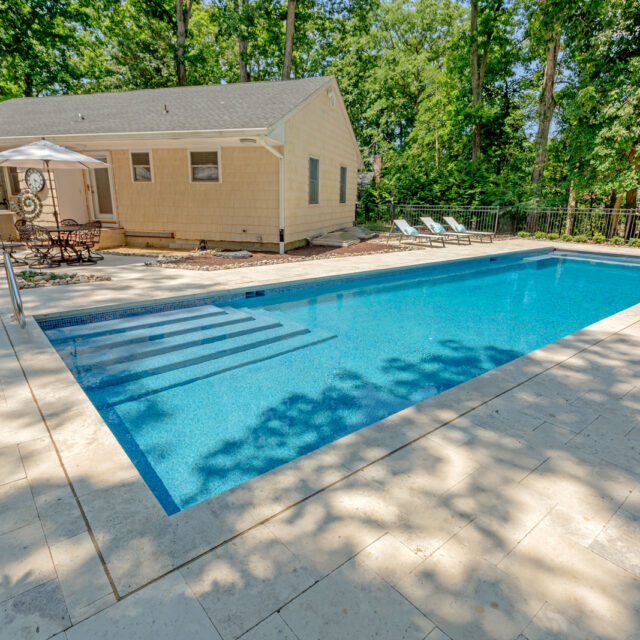 Rectangular inground pool with steps, surrounded by a stone patio, lounge chairs, and umbrellas next to a beige house and trees in the background—perfect for relaxing by luxury pools and enjoying outdoor kitchens.