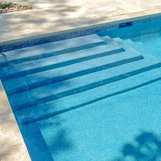 Wide steps lead into a clear blue inground pool with a metal handrail on a stone-tiled deck; some shadows from trees are visible on the water and deck.
