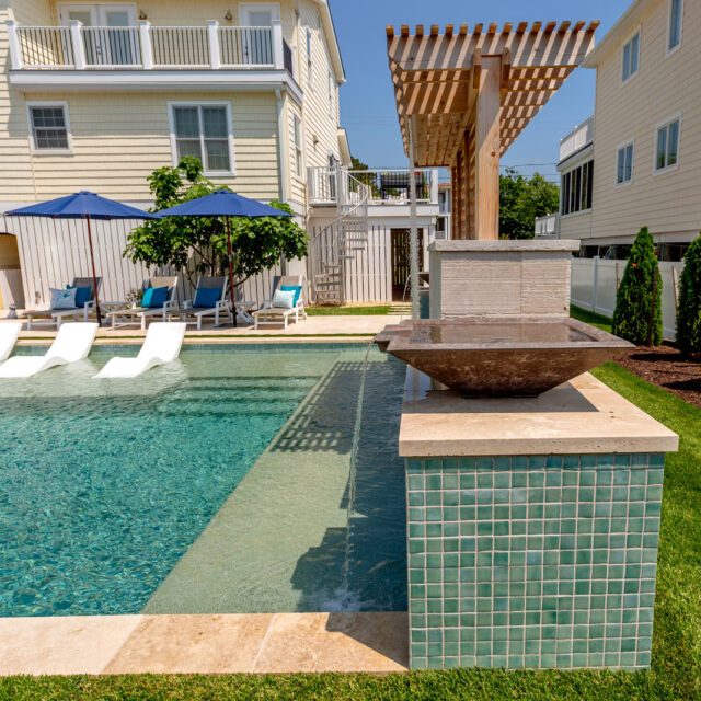 A backyard swimming pool with lounge chairs partially submerged in water, a water feature with green tiles, custom spas, and two beige houses in the background—perfect for elevated outdoor living.