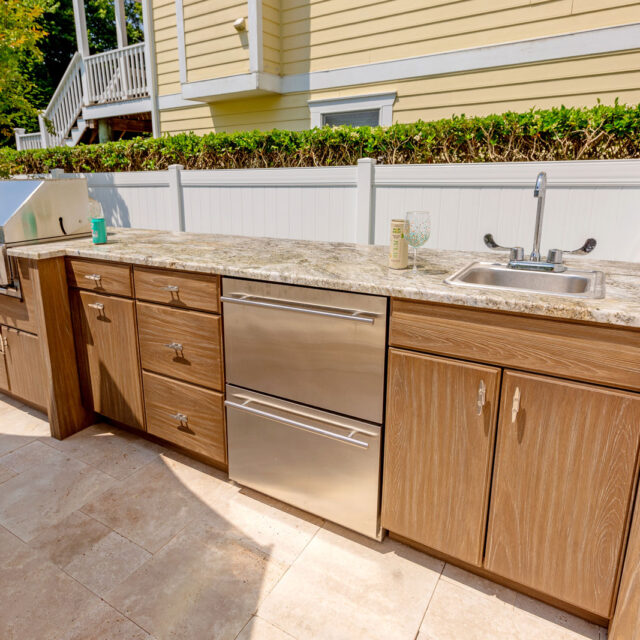 Outdoor kitchen with stainless steel grill, sink, drawers, and cabinets under a stone countertop on a tiled patio—perfect for outdoor living—set beside a white fence and green bushes.
