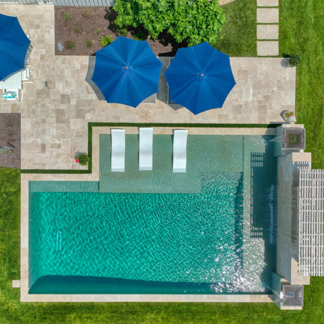 Aerial view of a luxury pool with three submerged lounge chairs and two blue umbrellas on a stone patio surrounded by green grass.