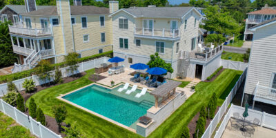 Aerial view of a large, two-story house with a fenced backyard featuring a luxury inground pool, lounge chairs, umbrellas, and manicured landscaping.