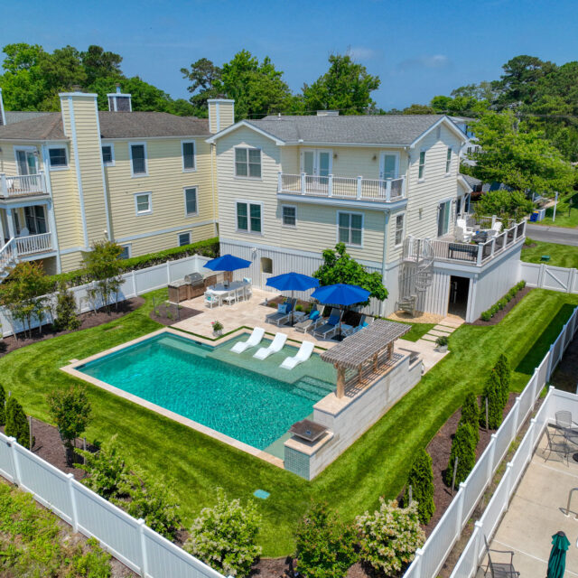 Aerial view of a large, two-story house with a fenced backyard featuring a luxury inground pool, lounge chairs, umbrellas, and manicured landscaping.