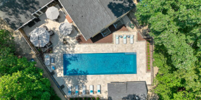 Aerial view of a rectangular luxury pool in the backyard, surrounded by lounge chairs, patio furniture, and trees next to a house—perfect for outdoor living.