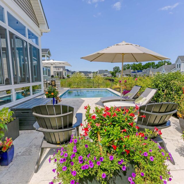 Poolside patio with lounge chairs, umbrella, and black chairs surrounds luxury pools, accented by colorful potted flowers and greenery on a sunny day in a residential neighborhood.