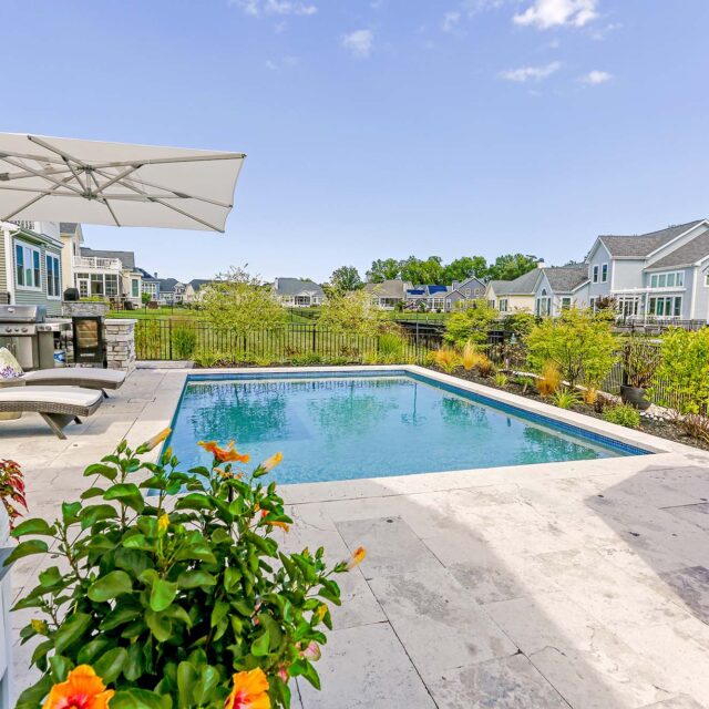 Private backyard with a rectangular inground pool, lounge chairs, patio umbrellas, potted plants, and a view of neighboring houses under a clear blue sky.