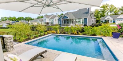 Rectangular luxury pool with two lounge chairs on a stone patio, surrounded by a black fence and landscaped garden—perfect for relaxing or entertaining near outdoor kitchens, with houses in the background.