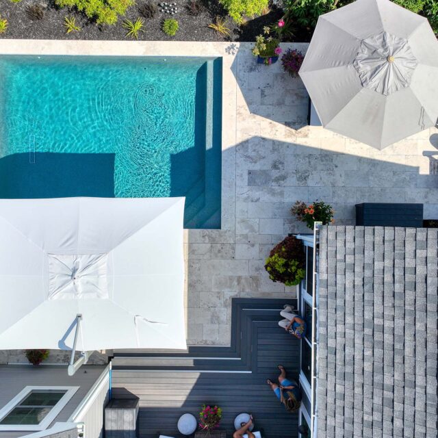 Aerial view of a backyard with a luxury pool, lounge chairs, umbrellas, a tiled patio for outdoor living, landscaped garden, and part of a house with people sitting on the deck.