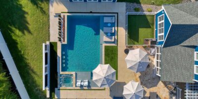Aerial view of a backyard with a rectangular inground pool, custom spa, lounge chairs, umbrellas, and patio area next to a house and outbuilding—perfect for enjoying luxury pools living.