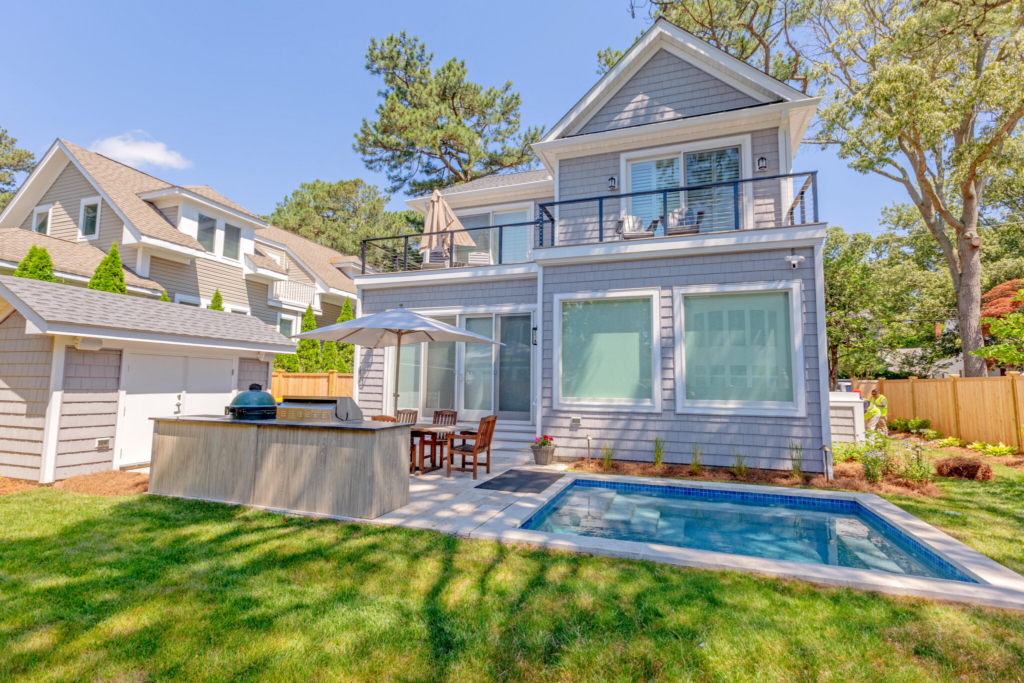 Modern backyard with a small plunge pool, outdoor kitchen, dining area, and a two-story gray house surrounded by trees and greenery.