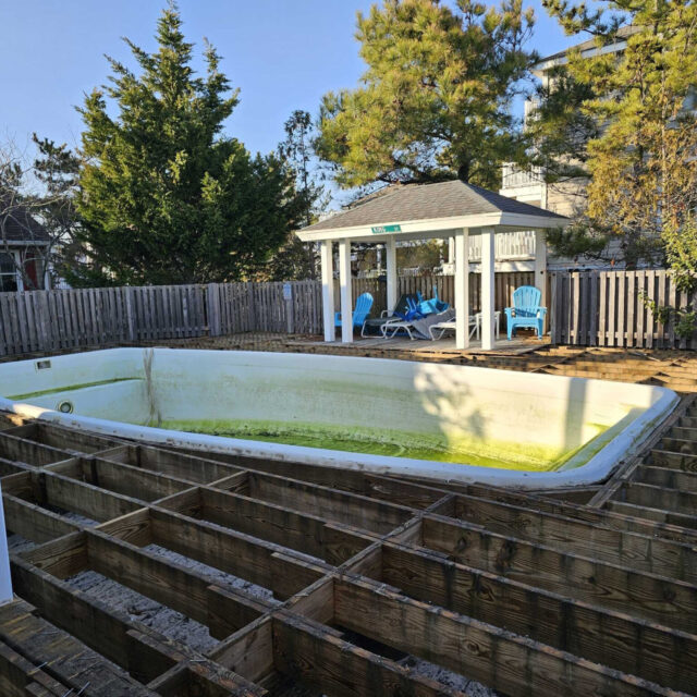 An empty inground pool with dirty water at the bottom sits in a backyard surrounded by a partially dismantled wooden deck, creating an outdoor living space with a gazebo and blue chairs in the background.