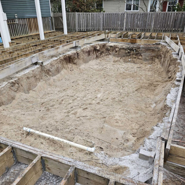 A construction site with a large, oval-shaped sand pit surrounded by wooden framing and exposed dirt, possibly the early stages of installing fiberglass pools or other types of inground pools.