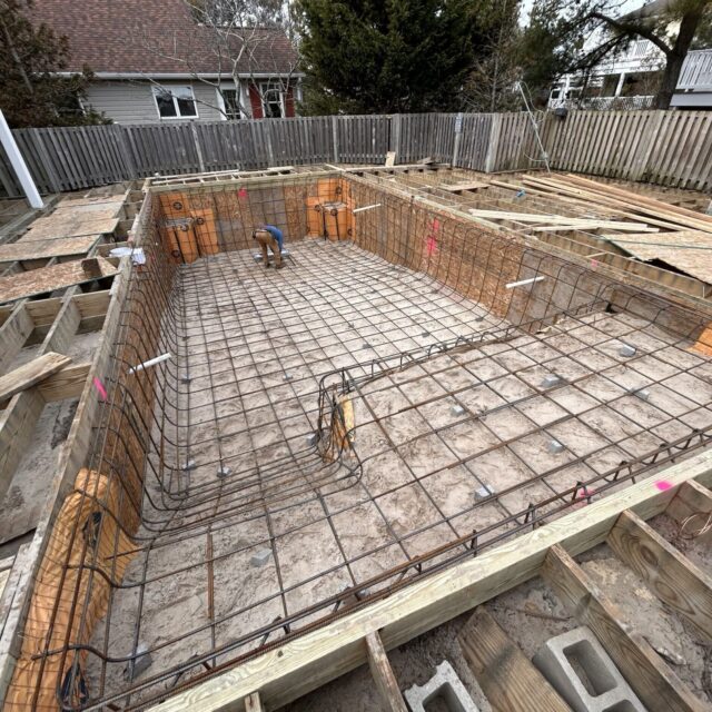 A rectangular luxury pool is under construction, featuring rebar framework and wooden forms, with a worker inside the pool area.