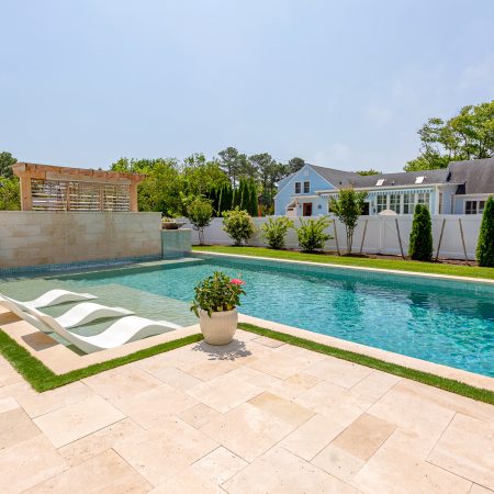 A rectangular backyard luxury pool with built-in lounge chairs, a water feature, and stone tile patio is surrounded by fencing and landscaping—perfect for transforming your space alongside custom spas or fiberglass pools.