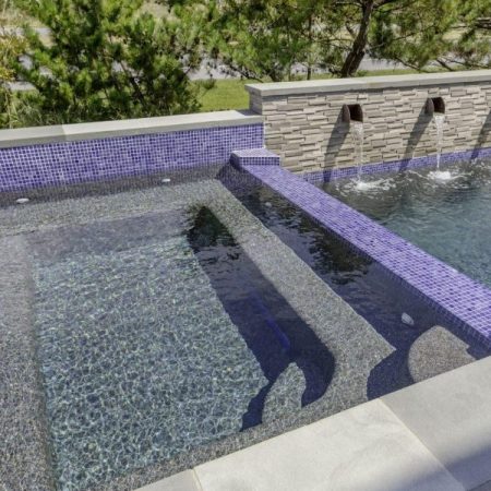 A modern backyard luxury pool with a raised custom spa section, purple tile accents, and water features flowing from a stone wall into the pool.