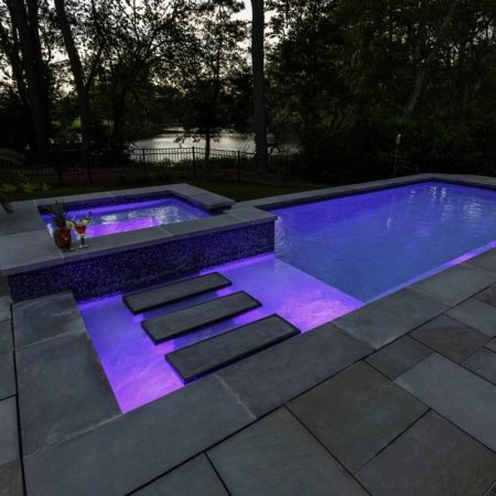 A modern backyard luxury pool and hot tub with purple underwater lights, surrounded by gray stone tiles, steps inside the pool, and trees in the background.