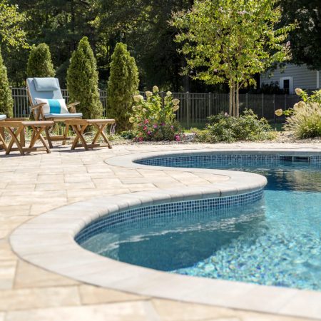 A backyard swimming pool with blue water, surrounded by a stone patio. Three cushioned lounge chairs and wooden tables line the fiberglass pool, creating an inviting outdoor living space with trees and plants in the background.