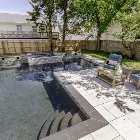 Rectangular luxury pool with built-in spa, water feature, and steps; two blue cushioned chairs and a table rest on a tiled patio surrounded by trees and a wooden fence—perfect for relaxing by one of the finest inground pools.