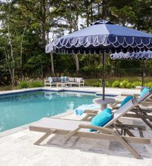 Outdoor pool area with luxury pools, lounge chairs, blue cushions, and large blue umbrellas, surrounded by trees and patio furniture on a stone deck.