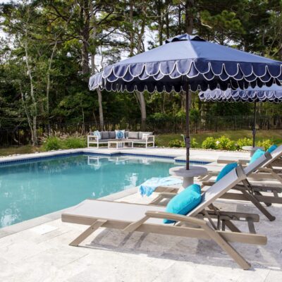 Outdoor pool area with luxury pools, lounge chairs, blue cushions, and large blue umbrellas, surrounded by trees and patio furniture on a stone deck.