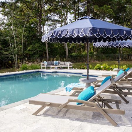 Outdoor pool area with luxury pools, lounge chairs, blue cushions, and large blue umbrellas, surrounded by trees and patio furniture on a stone deck.
