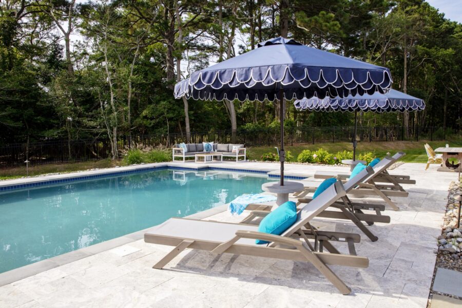 Outdoor pool area with luxury pools, lounge chairs, blue cushions, and large blue umbrellas, surrounded by trees and patio furniture on a stone deck.