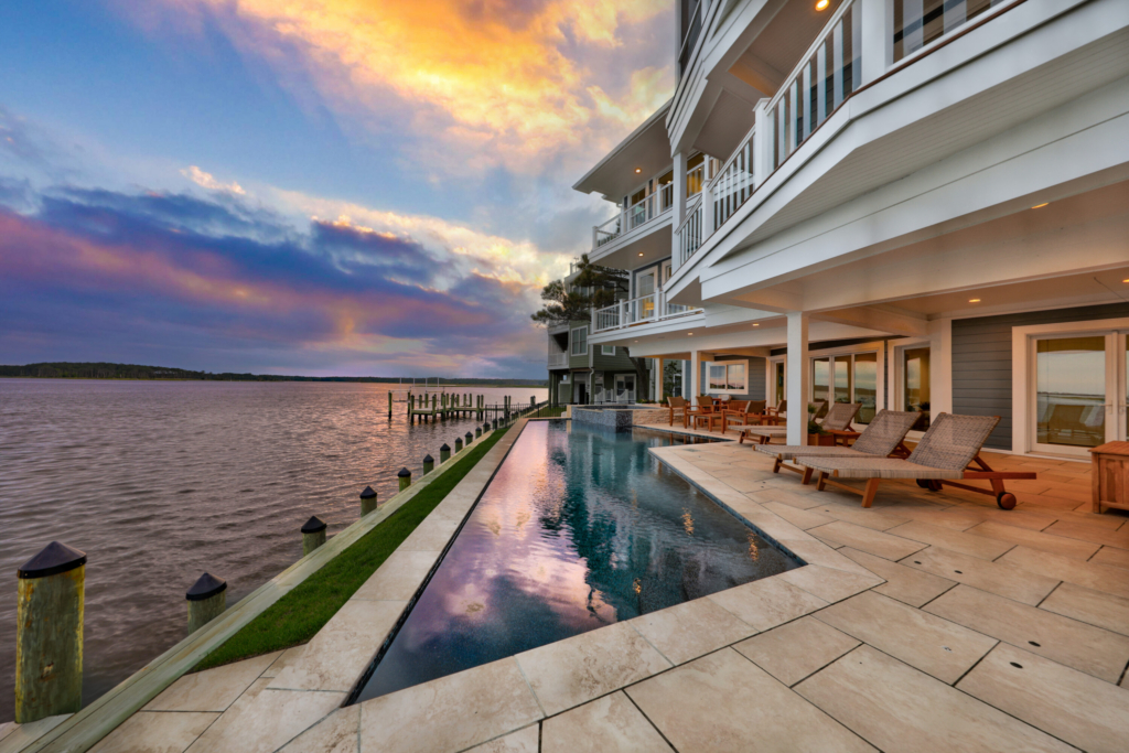 Modern waterfront house with multiple balconies, an infinity pool, and lounge chairs overlooking a calm body of water at sunset—perfect for outdoor living and relaxing in style.